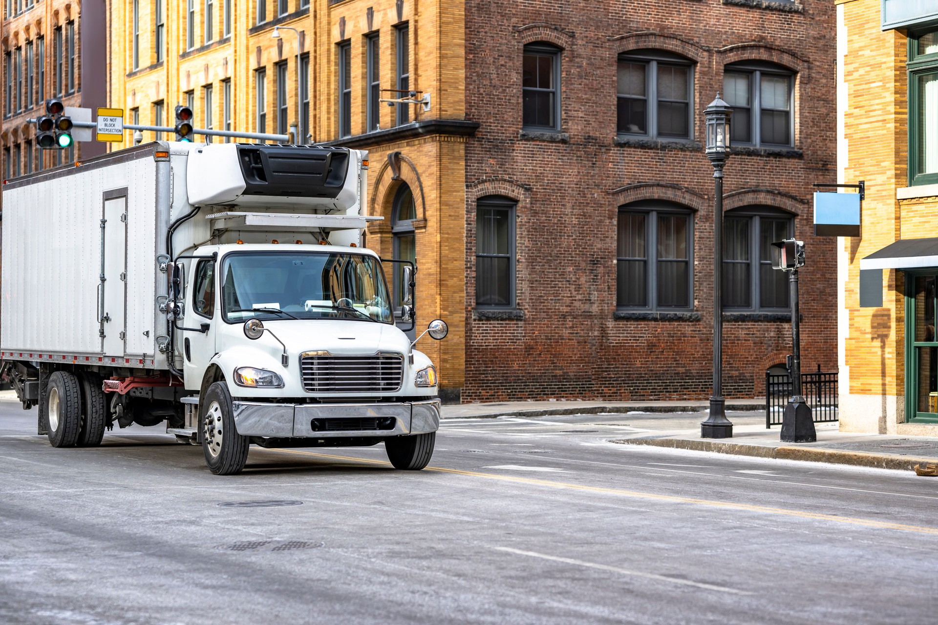 Day cab rig semi truck transporting cargo in long refrigerated box trailer driving on the crossroad intersection of the old Boston city street with bricks multilevel buildings
